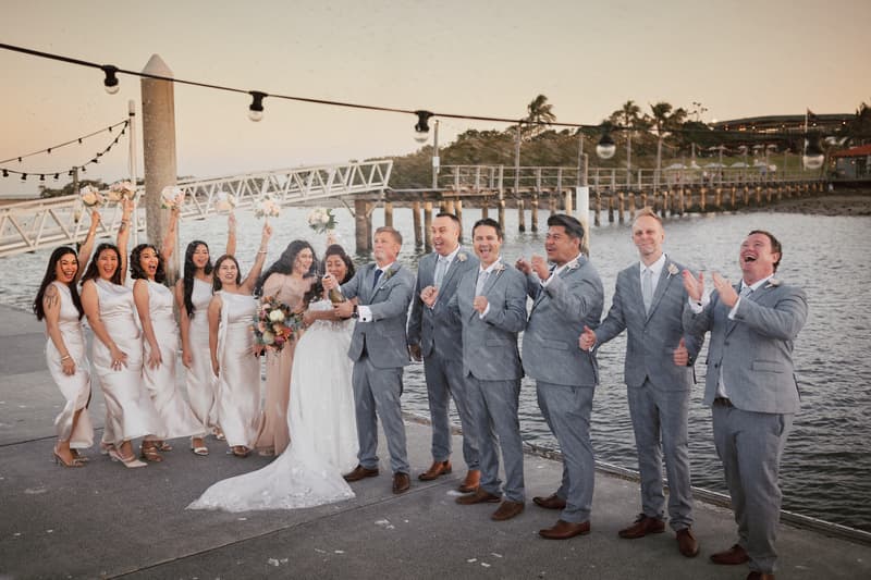 The bride Jheng and groom Dane stand on a waterfront pier at Sandstone Point Hotel with their bridal party. The bride is in a white wedding gown holding a bouquet, and the groom is in a grey suit. Bridesmaids in matching light-colored dresses raise their bouquets, and groomsmen in grey suits cheer as the groom pops a champagne bottle.