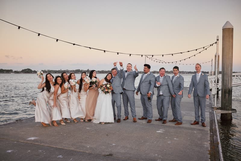 The bride Jheng in a white wedding gown and the groom Dane in a grey suit stand with their bridal party on a pier at Sandstone Point Hotel. The bridesmaids wear light-colored dresses and hold bouquets, while the groomsmen wear matching grey suits. String lights hang above them with water and distant trees in the background.