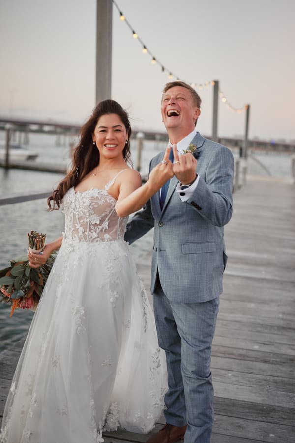 Bride Jheng and groom Dane stand on a wooden pier at Sandstone Point Hotel, smiling and showing their wedding rings to the camera. Jheng holds a bouquet and wears a white floral lace wedding gown, while Dane wears a light grey suit with a boutonniere.