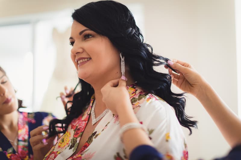The bride Kelly has her earrings and hair adjusted by attendants while wearing a floral robe, with a wedding dress blurred in the background at Novotel Sunshine Coast Resort.