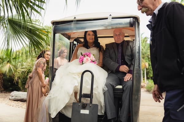 The bride Kelly sits in a golf cart holding a bouquet of pink and white flowers, accompanied by an older man in a suit, at Novotel Sunshine Coast Resort. Two bridesmaids in matching dresses stand nearby, and a man in dark clothing is partially visible on the right.
