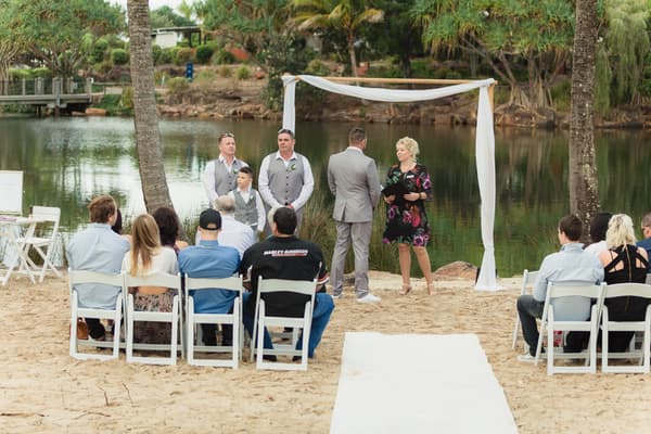 The groom Shane stands facing the celebrant under a simple white fabric-draped arch by a lake at Novotel Sunshine Coast Resort, with three groomsmen and a ring bearer standing nearby and guests seated on white chairs watching the ceremony.