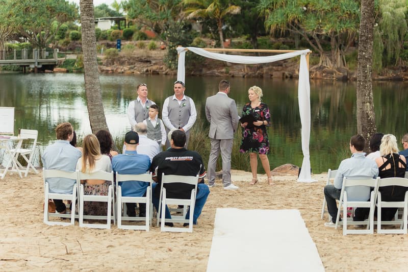 The groom Shane stands facing the celebrant under a simple white fabric-draped arch by a lake at Novotel Sunshine Coast Resort, with three groomsmen and a ring bearer standing nearby and guests seated on white chairs watching the ceremony.