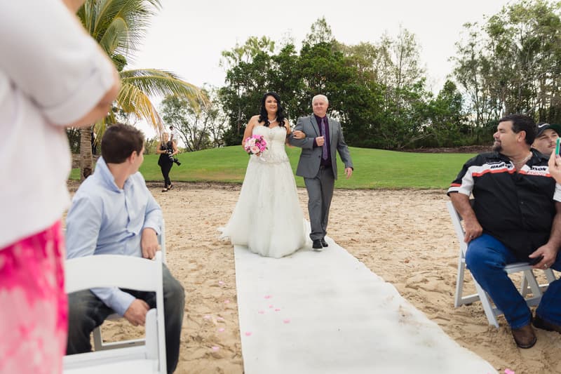 The bride Kelly walks arm-in-arm with an older man down a white aisle runner on sand at Novotel Sunshine Coast Resort, while seated guests watch.