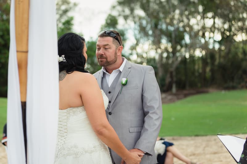 Kelly the bride and Shane the groom hold hands facing each other during their wedding ceremony at Novotel Sunshine Coast Resort, with greenery in the background.