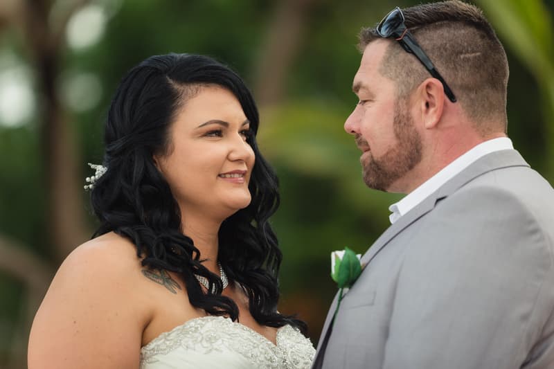 The bride Kelly and groom Shane face each other during their wedding ceremony at Novotel Sunshine Coast Resort, with Kelly in a strapless white gown and Shane in a light gray suit with a boutonniere.