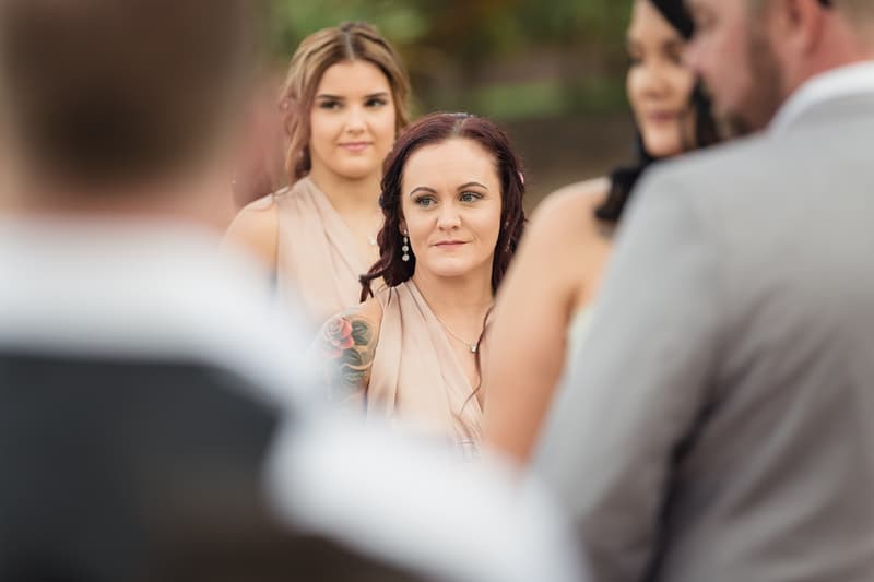 Two bridesmaids stand near the ceremony stage at Novotel Sunshine Coast Resort, with the bride and groom partially visible in the foreground.