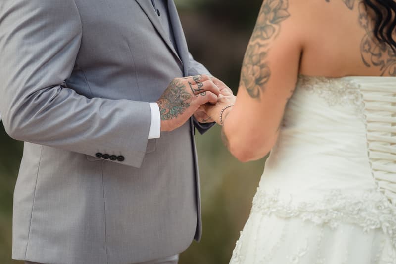 The groom in a light grey suit places a ring on the bride's finger, who is wearing a white strapless wedding dress with lace-up back and floral tattoo on her shoulder, at Novotel Sunshine Coast Resort.