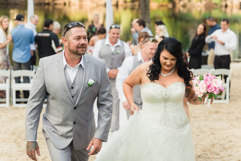 The bride Kelly in a white wedding gown holding a bouquet of pink and white flowers walks alongside the groom Shane in a light grey suit with a white boutonniere at Novotel Sunshine Coast Resort. Wedding guests and groomsmen in grey vests are visible in the background.