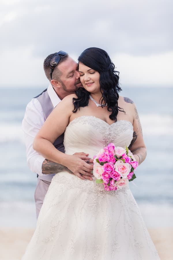 Bride Kelly in a white wedding gown holding a bouquet of pink and white flowers is embraced from behind by groom Shane wearing a light-colored vest and white shirt, with the ocean in the background.