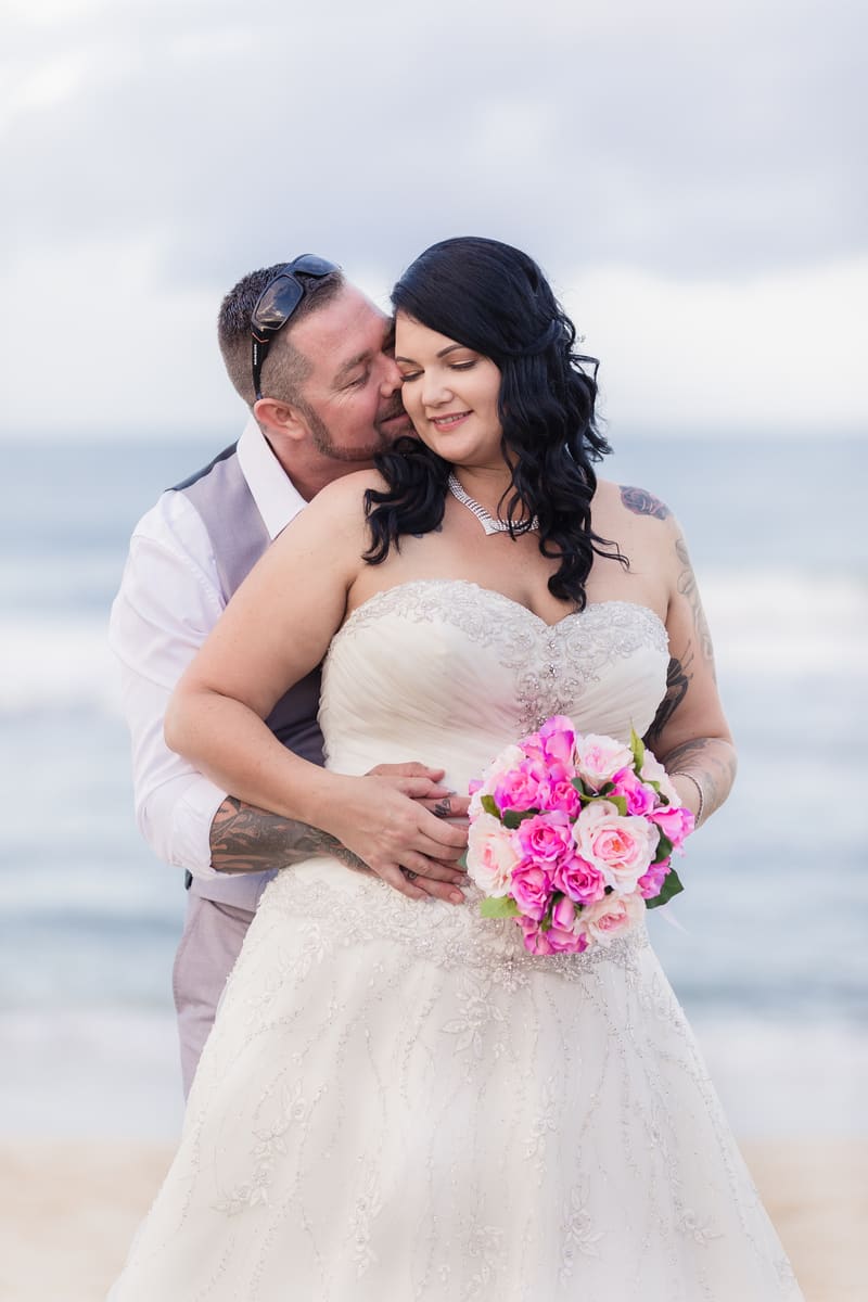 Bride Kelly in a white wedding gown holding a bouquet of pink and white flowers is embraced from behind by groom Shane wearing a light-colored vest and white shirt, with the ocean in the background.