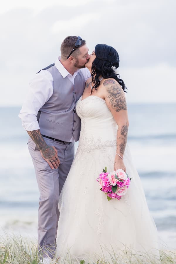 Kelly the bride and Shane the groom kiss outdoors near the water, with Kelly holding a bouquet of pink flowers.