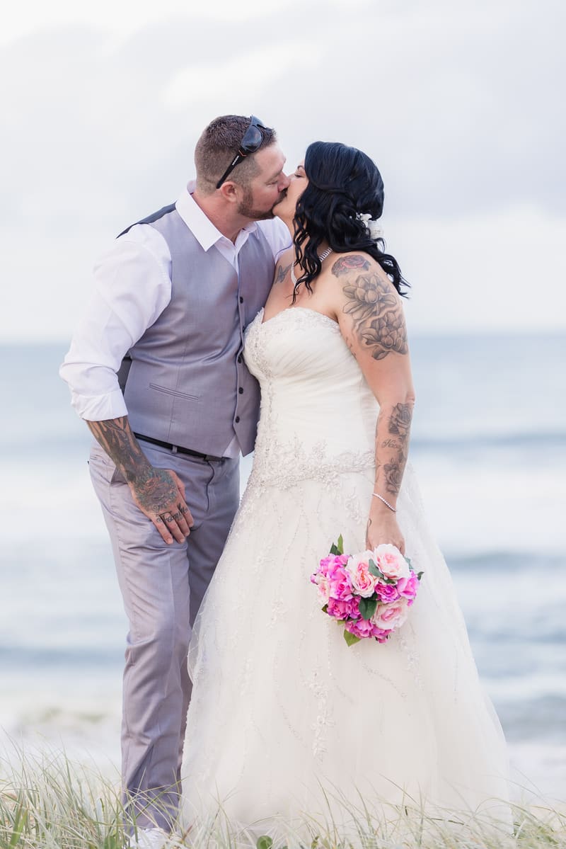 Kelly the bride and Shane the groom kiss outdoors near the water, with Kelly holding a bouquet of pink flowers. Brisbane videographer by She Said Yes.