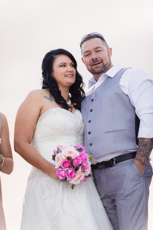 Kelly the bride in a white wedding gown holding a pink and white bouquet stands next to Shane the groom wearing a light grey vest and white shirt at Novotel Sunshine Coast Resort.