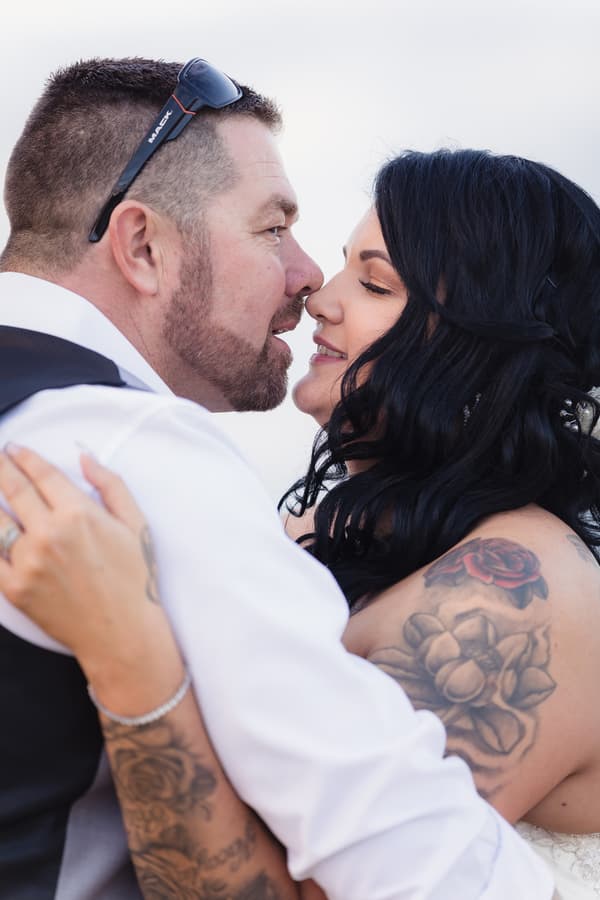 Bride Kelly and groom Shane embrace closely, about to kiss, showing their tattoos and wedding attire.