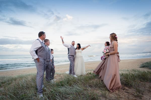 The bride Kelly and groom Shane pose with their bridal party on a beach with the ocean and sky in the background at Novotel Sunshine Coast Resort.