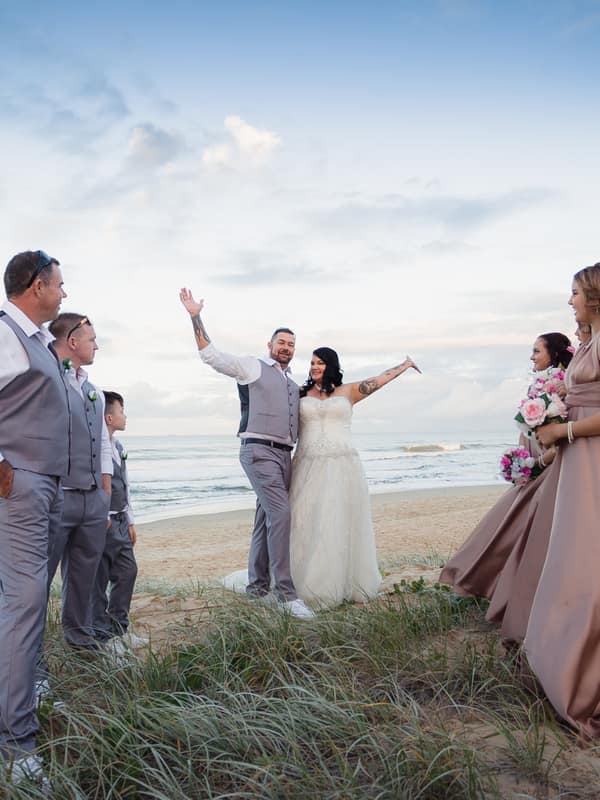 The bride Kelly and groom Shane pose with their bridal party on a beach with the ocean and sky in the background at Novotel Sunshine Coast Resort.