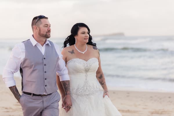 Kelly the bride and Shane the groom walk hand in hand on a beach with ocean waves in the background. Wedding photographers in Brisbane by She Said Yes.