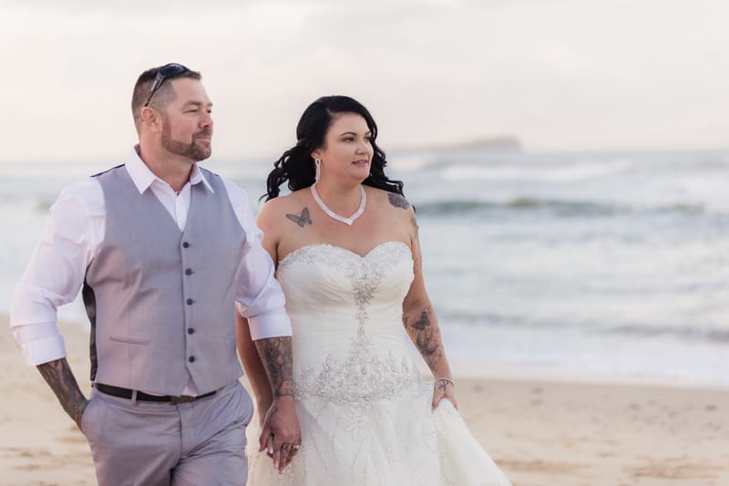 Kelly the bride and Shane the groom walk hand in hand on a beach with ocean waves in the background. Wedding photographers in Brisbane by She Said Yes.