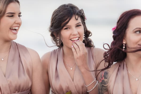 Three bridesmaids in matching mauve dresses stand together outdoors, with the central bridesmaid smiling and touching her lips. What is an engagement party by She Said Yes.