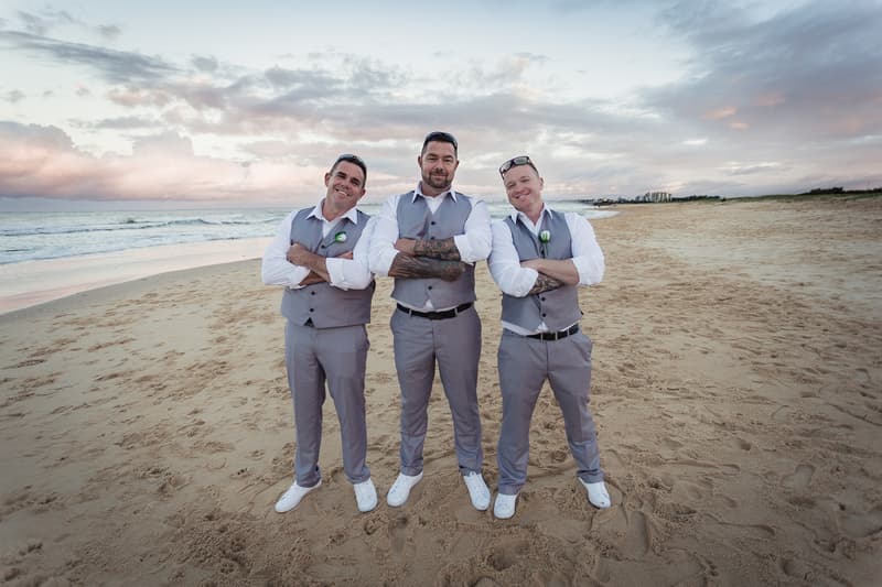 Three groomsmen stand on a sandy beach with arms crossed, wearing matching grey vests, white shirts, and white shoes, with the ocean and cloudy sky in the background.