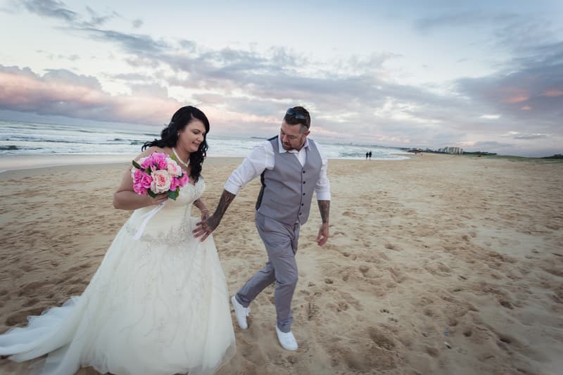 Kelly in her wedding dress holding a bouquet and Shane in a grey vest and pants walk together on a sandy beach near the ocean at sunset.