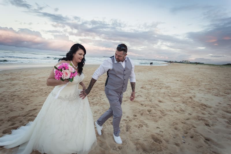 Kelly in her wedding dress holding a bouquet and Shane in a grey vest and pants walk together on a sandy beach near the ocean at sunset. Candid wedding photographer by She Said Yes.
