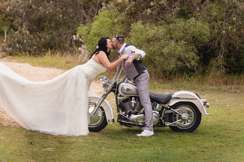 The bride Kelly in a white wedding gown leans forward to kiss the groom Shane who is standing next to a silver motorcycle on grass with trees in the background at Novotel Sunshine Coast Resort.