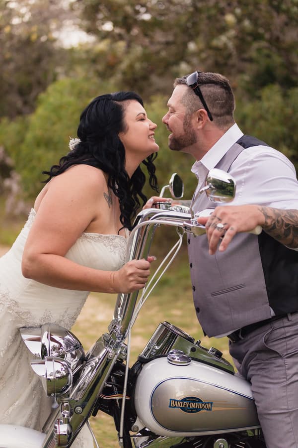 The bride Kelly and groom Shane lean toward each other over a Harley-Davidson motorcycle outdoors at Novotel Sunshine Coast Resort.