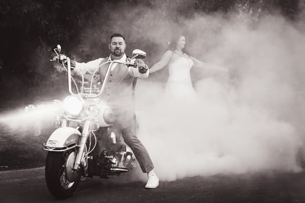 The groom rides a motorcycle with its headlights on, creating smoke around the bride who stands behind him with arms outstretched. Engagement parties by She Said Yes.
