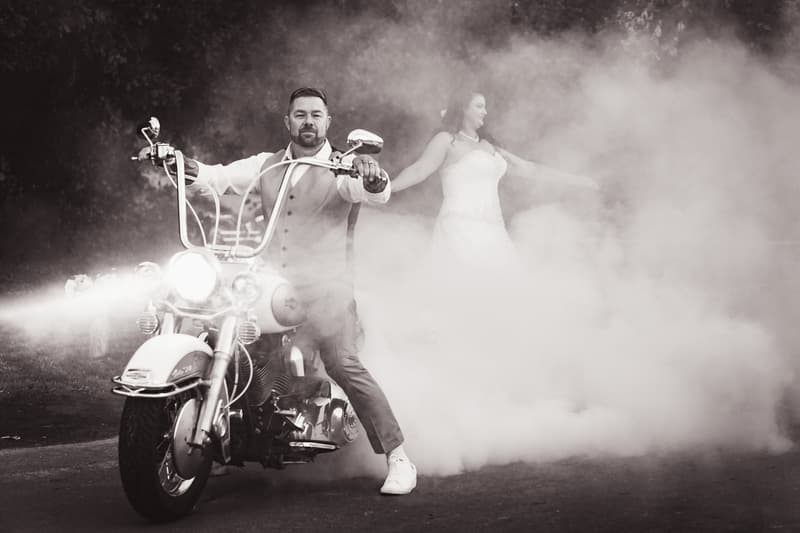 The groom rides a motorcycle with its headlights on, creating smoke around the bride who stands behind him with arms outstretched at Novotel Sunshine Coast Resort.