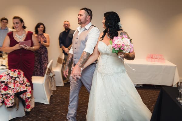 Bride Kelly in a white wedding gown holding a bouquet of pink and white flowers walks hand-in-hand with groom Shane, who is wearing a light grey vest and white shirt, at the reception stage of Novotel Sunshine Coast Resort. Guests stand and watch in the background.