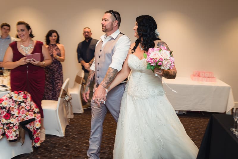 Bride Kelly in a white wedding gown holding a bouquet of pink and white flowers walks hand-in-hand with groom Shane, who is wearing a light grey vest and white shirt, at the reception stage of Novotel Sunshine Coast Resort. Guests stand and watch in the background.
