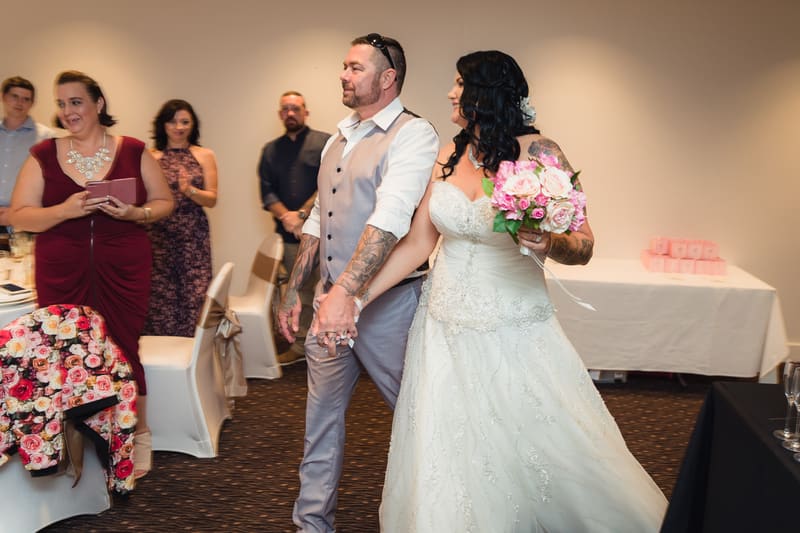 Bride Kelly in a white wedding gown holding a bouquet of pink and white flowers walks hand-in-hand with groom Shane, who is wearing a light grey vest and white shirt, at the reception stage of. Guests stand and watch in the background. Easter long weekend wedding Brisbane by She Said Yes.