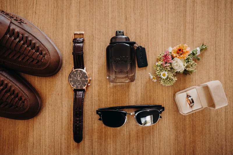 Groom's wedding accessories including brown woven shoes, a wristwatch with a leather strap, a bottle of cologne, black sunglasses, a small floral boutonniere, and a ring box containing two wedding bands arranged on a wooden surface.