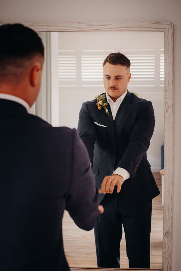 The groom Brandon adjusts his suit jacket while looking at himself in a large mirror in a room with white walls and wooden flooring at Sandstone Point Hotel.