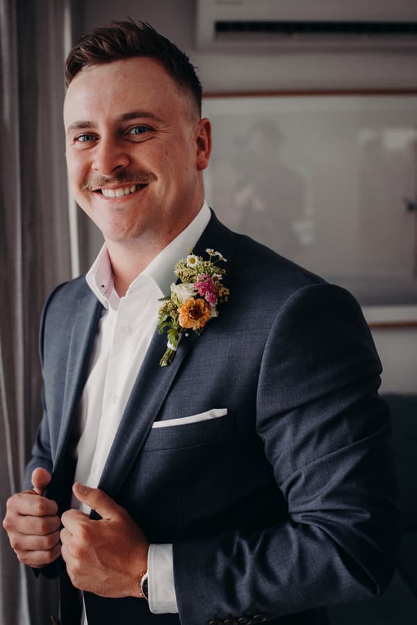 Brandon, the groom, adjusts his dark suit jacket indoors at Sandstone Point Hotel.