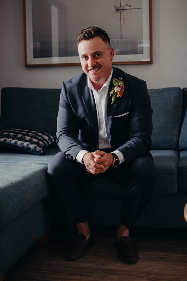 The groom Brandon sits on a blue couch wearing a dark suit with a boutonniere, smiling at the camera inside a room at Sandstone Point Hotel.