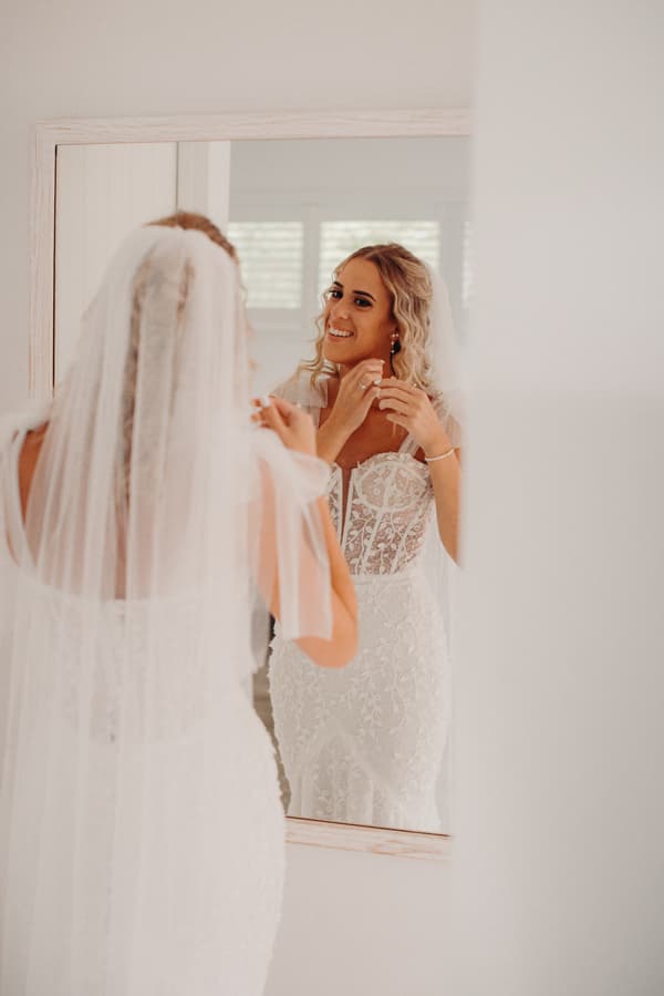The bride Krystal adjusts her earrings while looking at herself in a mirror indoors.