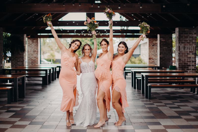The bride Krystal poses with three bridesmaids in peach dresses holding bouquets aloft inside the Sandstone Point Hotel Pavilion.