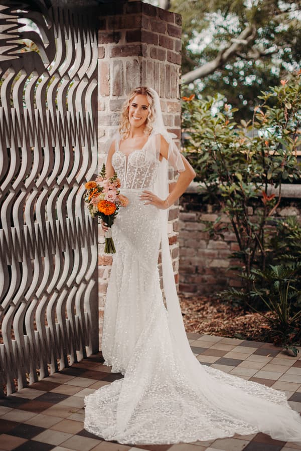 The bride Krystal stands alone holding a bouquet of flowers, posing against a brick pillar and decorative metal screen at Sandstone Point Hotel.