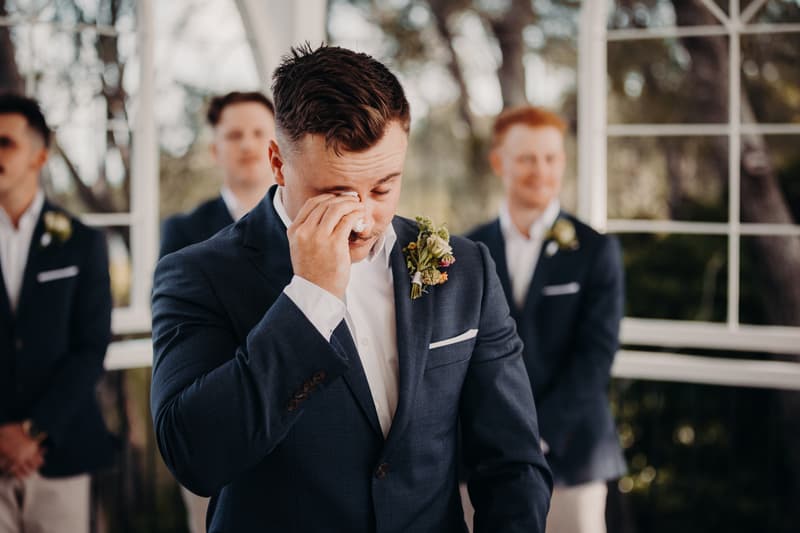 The groom wipes his eye with a tissue while standing at the ceremony stage at Sandstone Point Hotel — Pavilion, with groomsmen standing behind him.