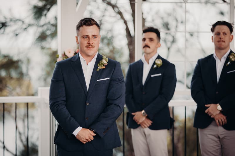 The groom and two groomsmen stand at the ceremony stage at Sandstone Point Hotel — Pavilion, all wearing navy blazers with boutonnieres and light-colored pants.