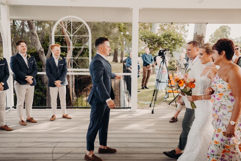 The bride Krystal, escorted by her parents, walks down the aisle towards the groom Brandon and groomsmen at the Sandstone Point Hotel — Pavilion during the ceremony.