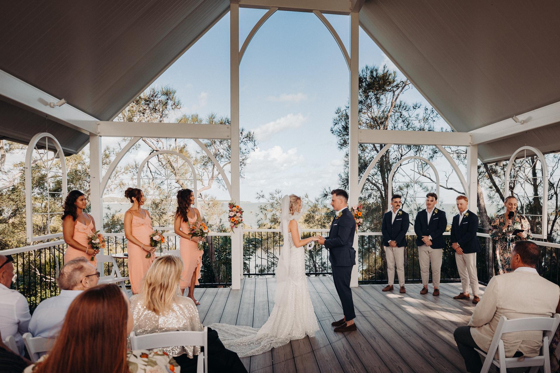 Bride Krystal and groom Brandon stand facing each other holding hands on the ceremony stage at Sandstone Point Hotel — Pavilion, surrounded by bridesmaids in peach dresses holding bouquets and groomsmen in navy blazers and beige pants, with guests seated in white chairs watching the ceremony.