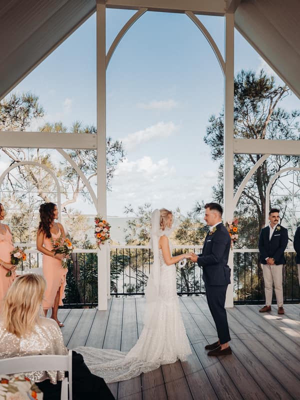 Bride Krystal and groom Brandon stand facing each other holding hands on the ceremony stage at Sandstone Point Hotel — Pavilion, surrounded by bridesmaids in peach dresses holding bouquets and groomsmen in navy blazers and beige pants, with guests seated in white chairs watching the ceremony.