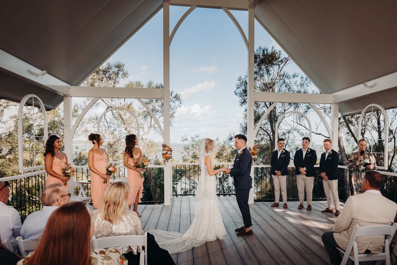 Bride Krystal and groom Brandon stand facing each other holding hands on the ceremony stage at Sandstone Point Hotel — Pavilion, surrounded by bridesmaids in peach dresses holding bouquets and groomsmen in navy blazers and beige pants, with guests seated in white chairs watching the ceremony.