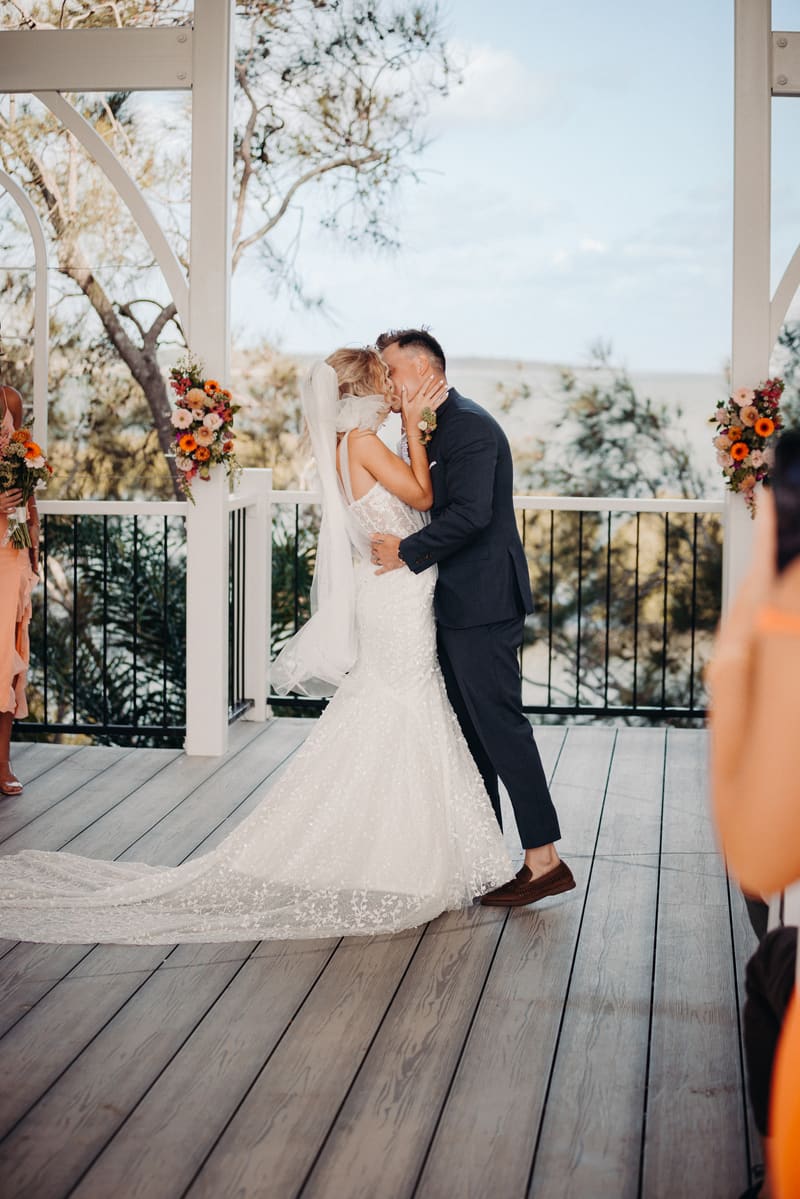 Bride Krystal and groom Brandon share a kiss on the ceremony stage at Sandstone Point Hotel — Pavilion, surrounded by floral arrangements and guests.