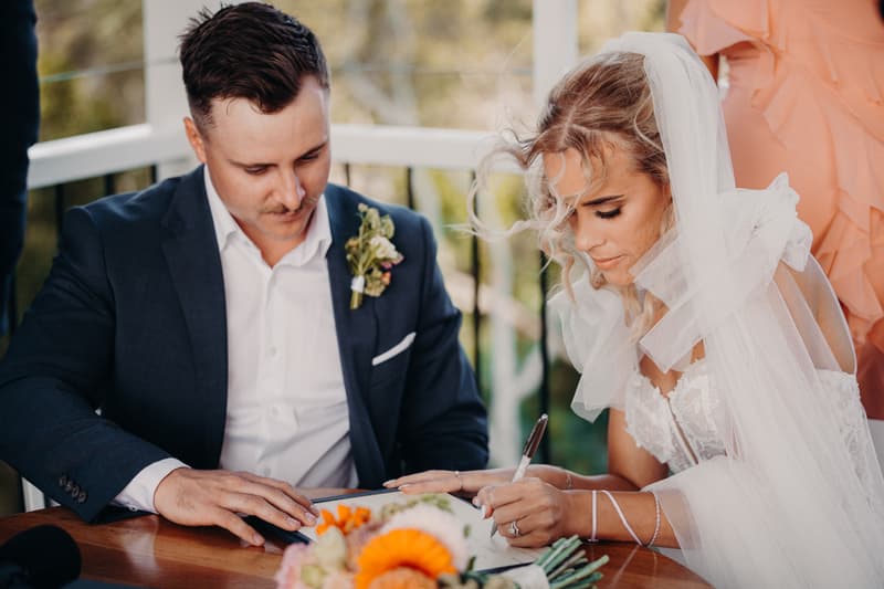 Bride Krystal and groom Brandon sit at a wooden table signing a document during their ceremony at Sandstone Point Hotel — Pavilion.