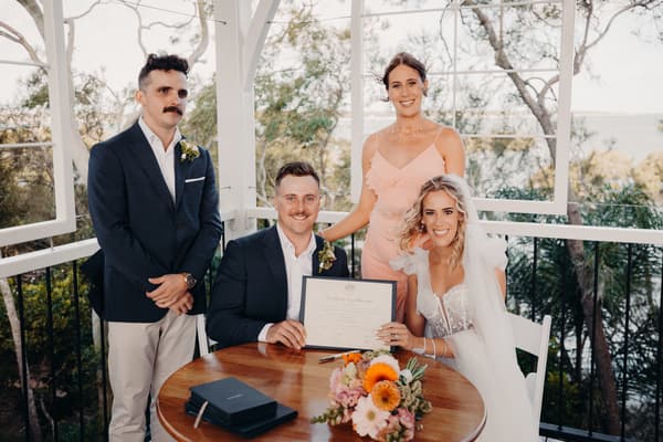 Krystal the bride and Brandon the groom sit at a round wooden table holding their marriage certificate at Sandstone Point Hotel — Pavilion, accompanied by a bridesmaid in a pink dress standing behind them and a groomsman in a navy blazer and beige pants standing beside them.